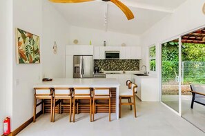A modern kitchen with white cabinets, a central island with bar stools, and a large window offering a view of greenery. - 3-Bedroom Jungle Villa with Pool & FirePit in lovely Puerto Viejo (Puerto Viejo de talamanca)