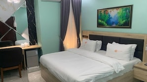 A hotel room with a bed, a desk, a chair, a nightstand, a painting, and a window with curtains. - P1 Place Hotel (Lagos)
