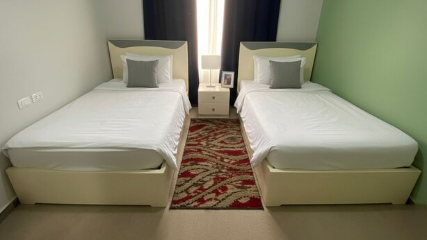 Quarto duplo com duas camas, tapete vermelho estampado, mesa de cabeceira com abajur e janela com cortinas.