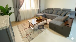 Uma sala de estar moderna com um sofá seccional, uma mesa de centro com frutas e petiscos, um tapete estampado e uma janela com cortinas.