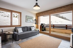 A modern living room with a grey sofa, a coffee table, and a large window with a view of a snowy landscape. - Chalet Angelus C201 - Two Bedroom Apartment, Sleeps 5 (Saint-Jean-de-Sixt)