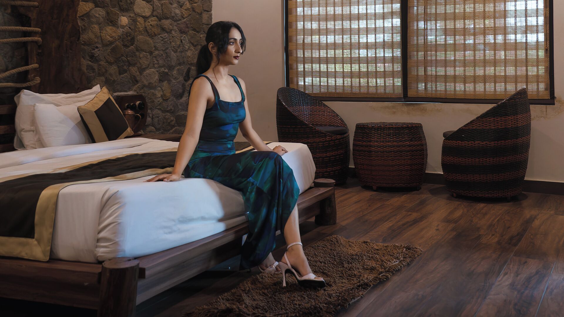 A room with a bed, a stone wall, a window with bamboo blinds, and two woven poufs.