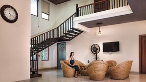 A spacious living area with a staircase, wicker furniture, a flat-screen TV, and a clock on the wall. - Corbett Mystic Inn An ExclusiveBoutique  (Ramnagar)