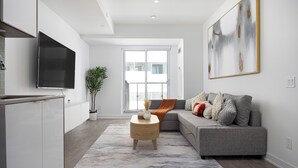 A modern living room with a grey sofa, a flat-screen TV mounted on the wall, a round wooden coffee table, and a large abstract painting.