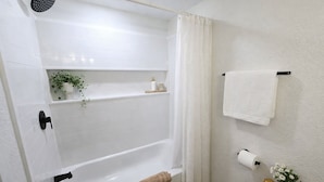 Un baño con una bañera blanca, ducha, toallero y estante con artículos de aseo.