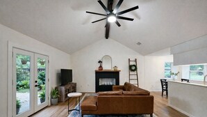 Un amplio salón con sofá de cuero marrón, chimenea, ventilador de techo y vistas al exterior a través de grandes puertas correderas de vidrio.