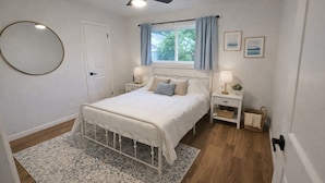Un dormitorio con cama, mesitas de noche, ventilador de techo, ventana con cortinas y espejo adosado a la pared.
