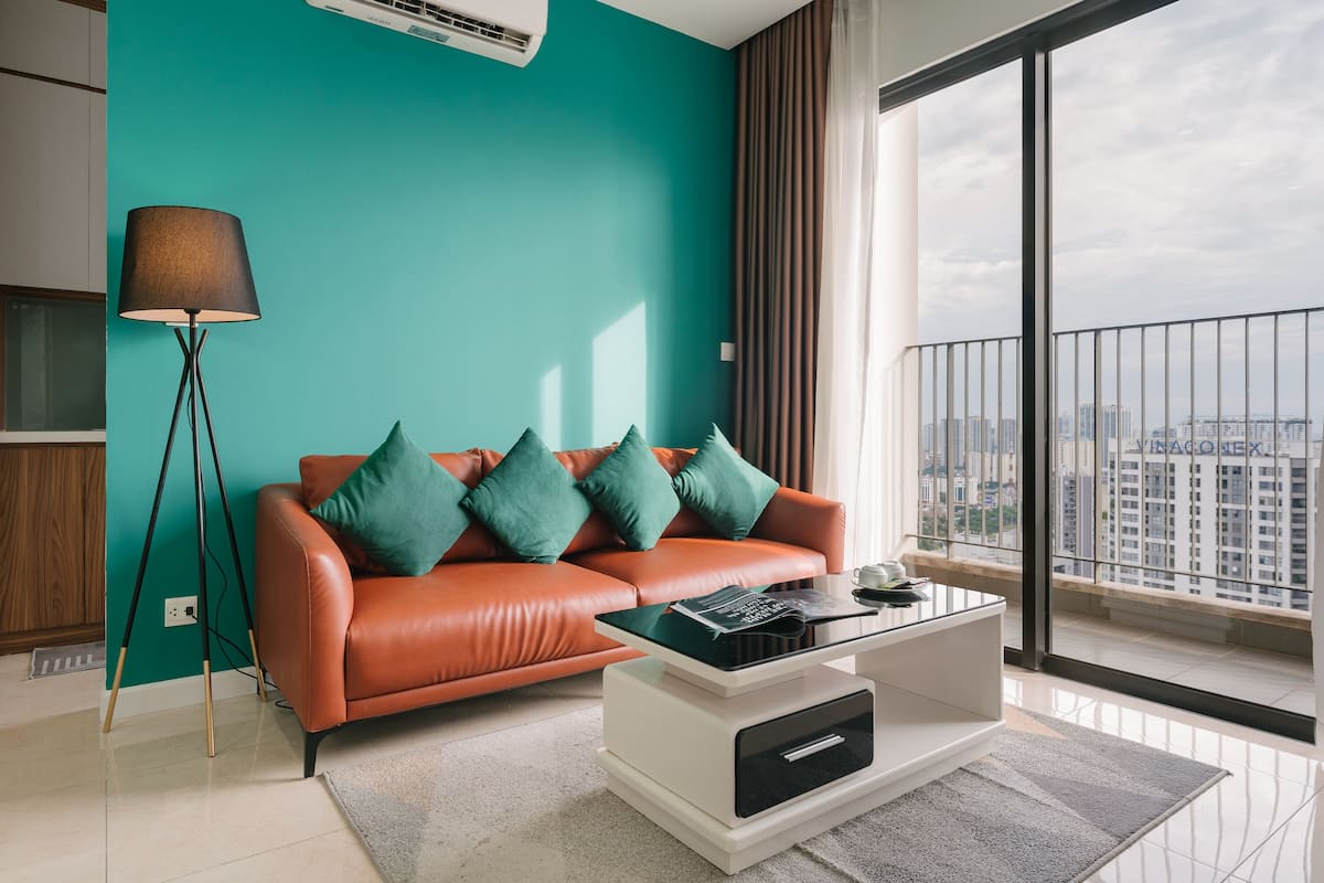 Un salon moderne avec un mur d’accent de couleur turquoise, un canapé en cuir agrémenté de coussins verts, une table basse en verre et une vue sur la ville grâce à de grandes fenêtres.