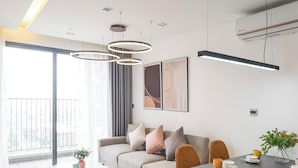 A modern living room with a sofa, dining table, and chairs, large windows, and a ceiling light fixture. - Vinhomes D'Capital - Great Vacation (Hanoi)