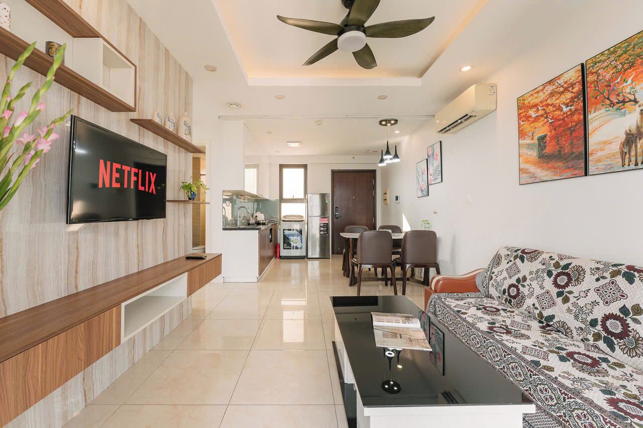 A modern living room with a flat-screen TV, a ceiling fan, a sofa, a coffee table, and a dining area with chairs.