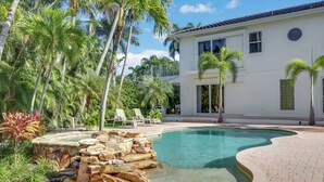 Une piscine bordée de pierres, des palmiers et un bâtiment blanc doté de fenêtres.