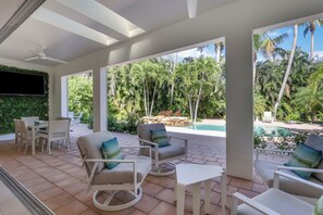 A covered outdoor seating area with chairs and a table, overlooking a pool and lush greenery. - EverHouse at the Moorings (Naples)