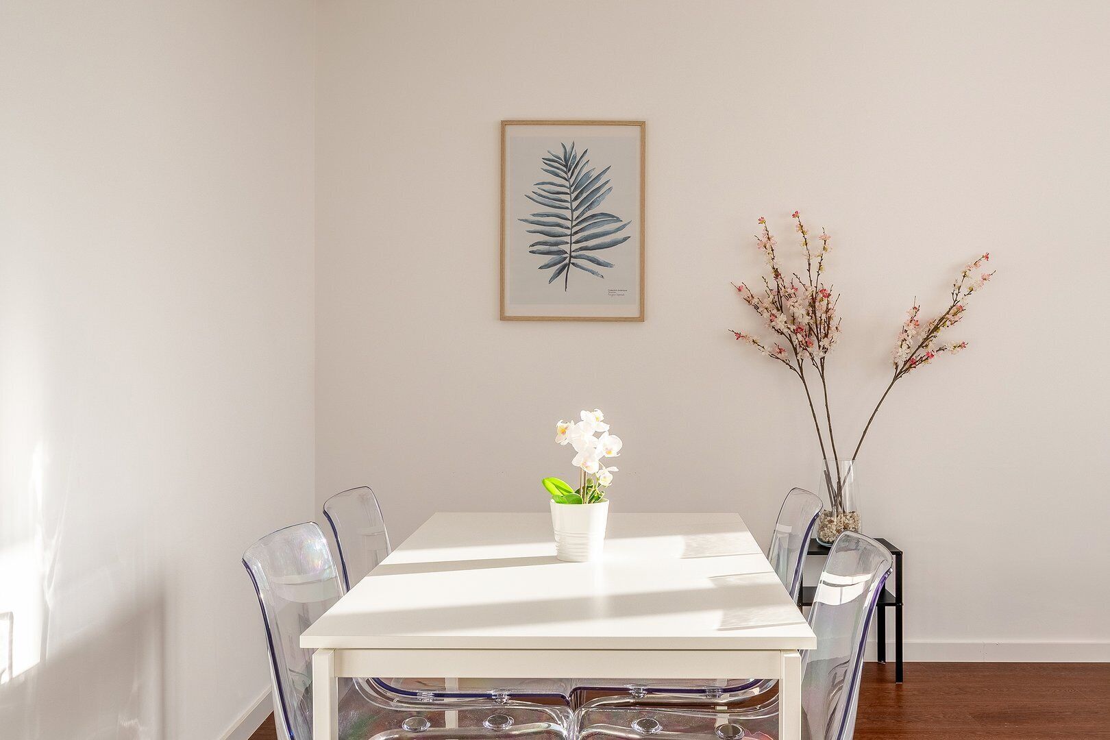 A dining area with a white table, clear chairs, a framed botanical print on the wall, and a vase with flowers.