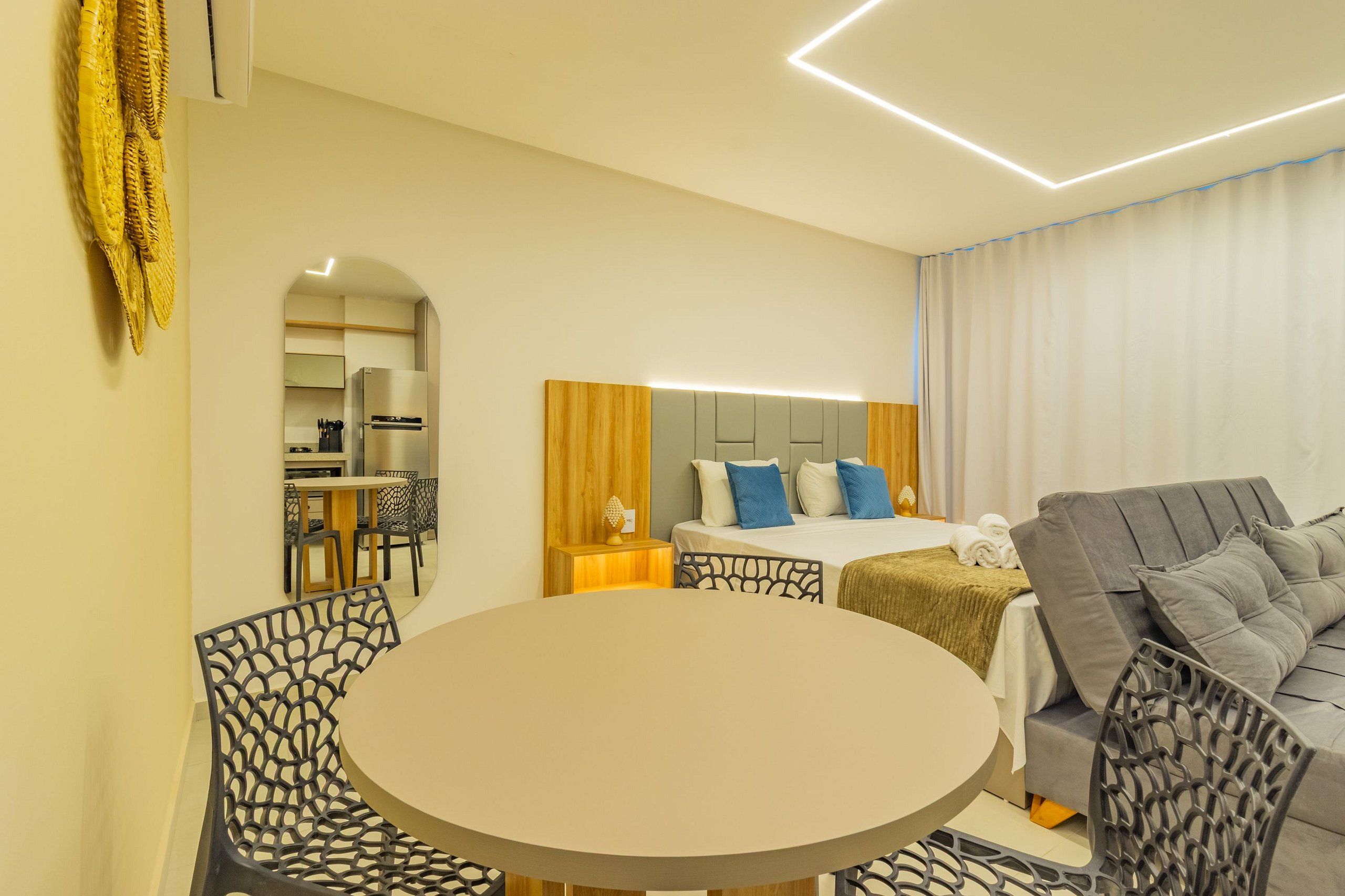 Quarto de hotel moderno com uma mesa de jantar redonda, um sofá, uma cama com roupa de cama azul e branca e uma cabeceira de madeira.