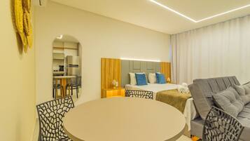 Quarto de hotel moderno com uma mesa de jantar redonda, um sofá, uma cama com roupa de cama azul e branca e uma cabeceira de madeira.