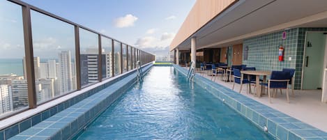 Una piscina infinita en la azotea con vistas a la ciudad, un área de descanso con mesas y sillas, y un piso de baldosas.