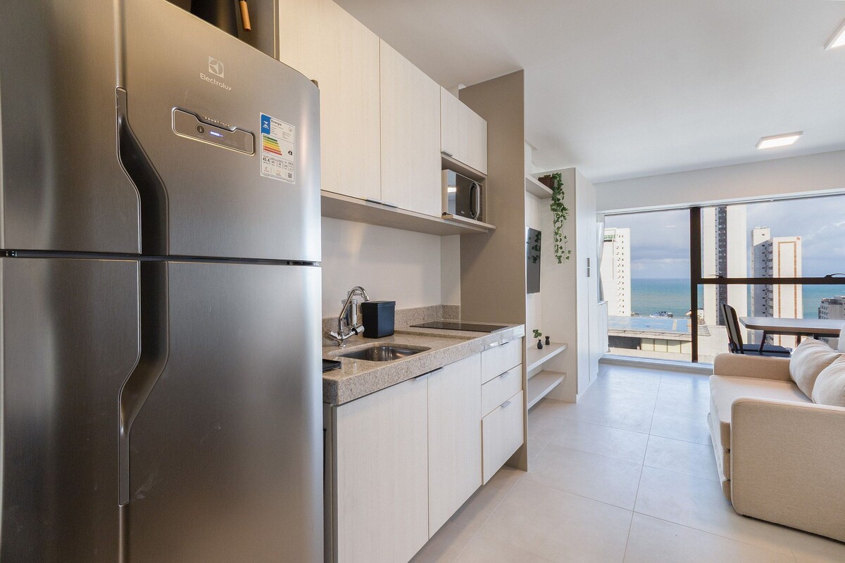 A modern kitchen with a stainless steel refrigerator, a double sink, and a microwave. There is a dining area with a table and chairs, and a view of the city through large windows.