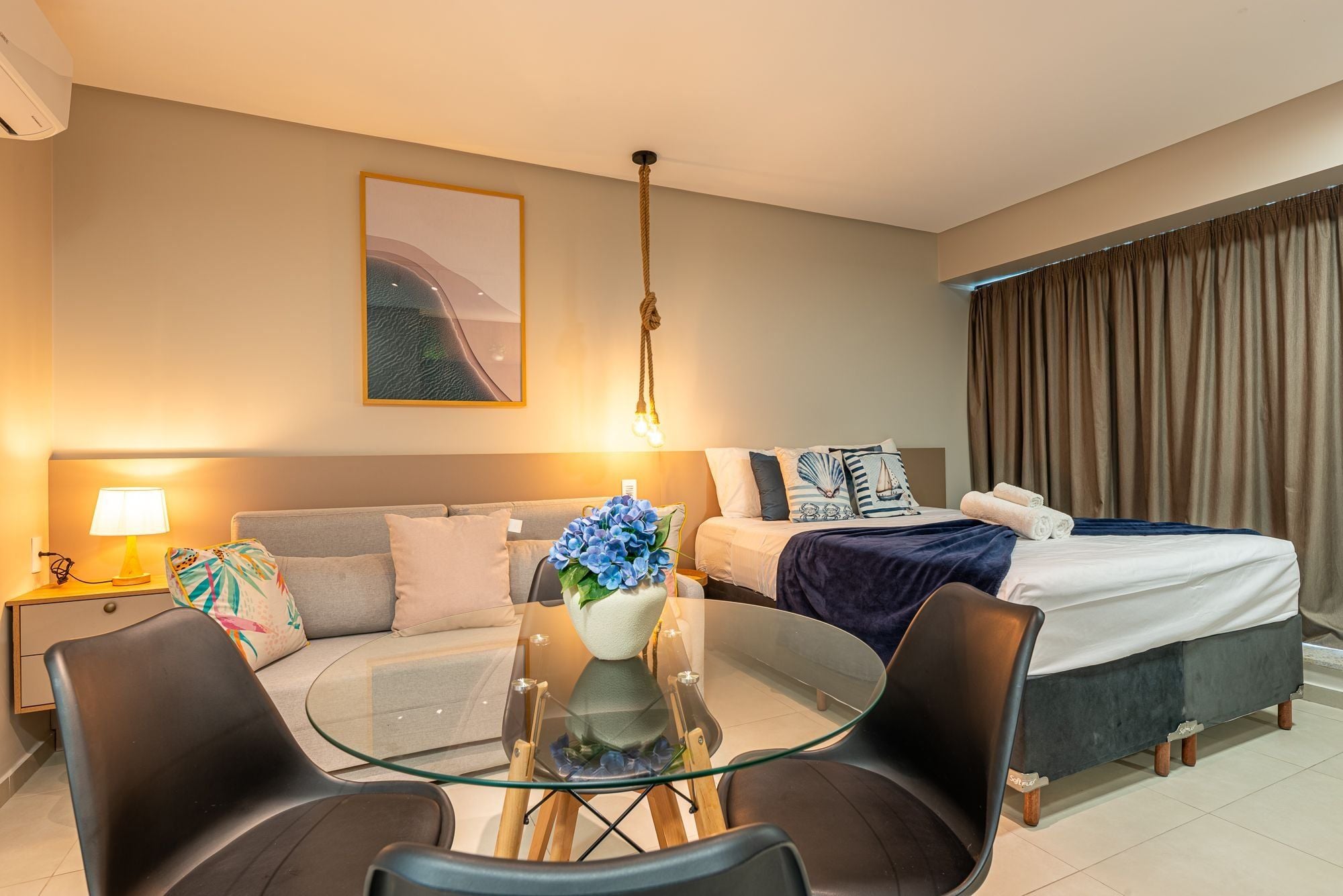 Habitación de hotel con cama, sofá, mesa de vidrio con sillas, lámpara y un cuadro en la pared.