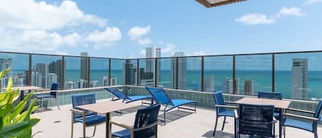 A rooftop terrace with a city view, tables, chairs, and potted plants.