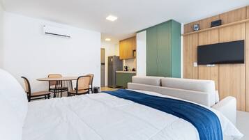 Quarto de hotel com uma cama grande, sofá, mesa de jantar com cadeiras, cozinha e televisão de tela plana na parede.