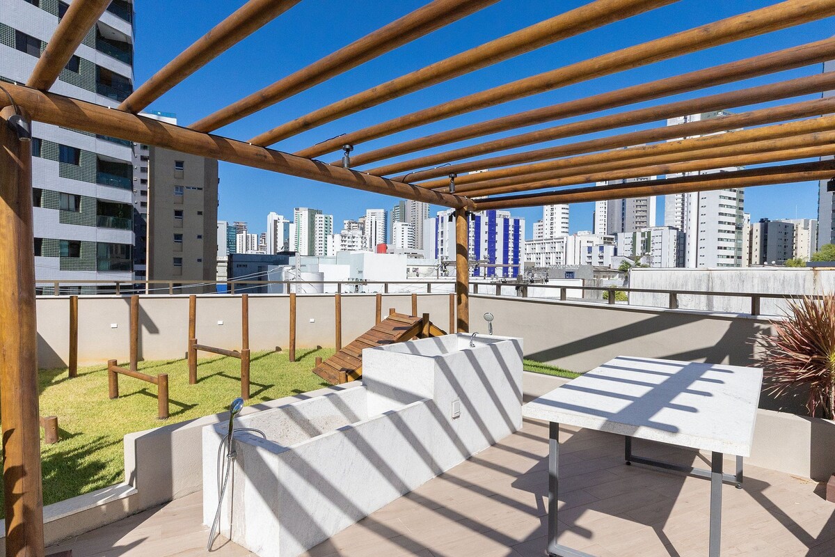 Een terras buiten met uitzicht op een stadsachtergrond, met een houten pergola, een witte tafel en een zithoek.