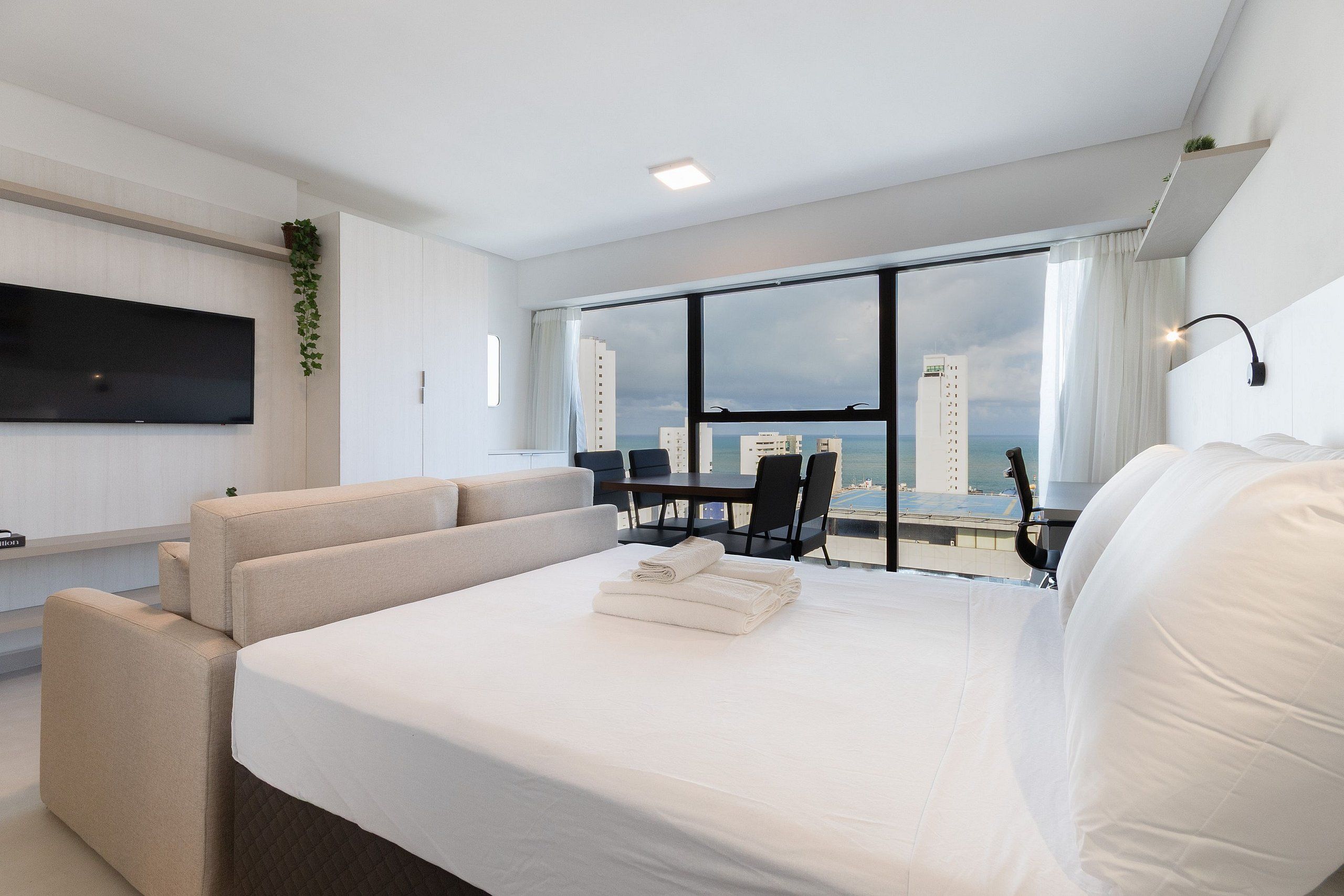 Una habitación de hotel moderna con una cama grande, un televisor de pantalla plana, una zona de comedor con mesa y sillas, y vistas a la ciudad y al océano.