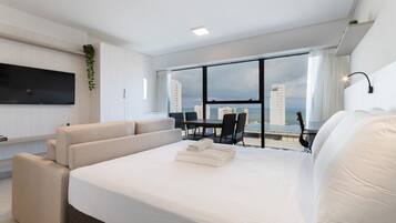 Una habitación de hotel moderna con una cama grande, un televisor de pantalla plana, una zona de comedor con mesa y sillas, y vistas a la ciudad y al océano.