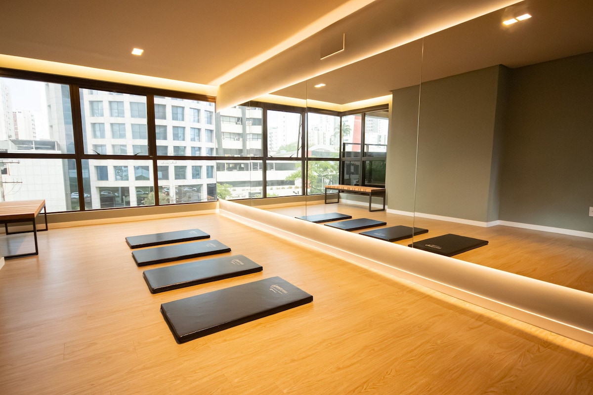 A modern gym with wooden flooring, exercise mats, and large windows offering city views.