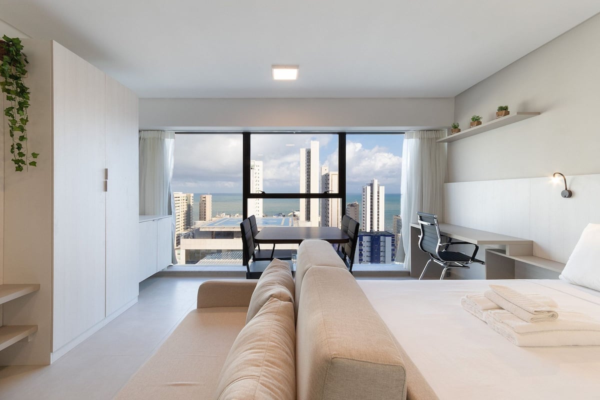 A modern hotel room with a large window, a sofa, a desk, and a chair.