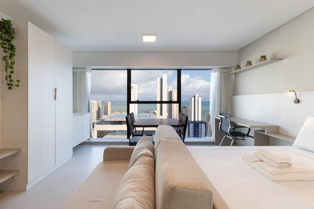 A modern hotel room with a large window, a sofa, a desk, and a chair.