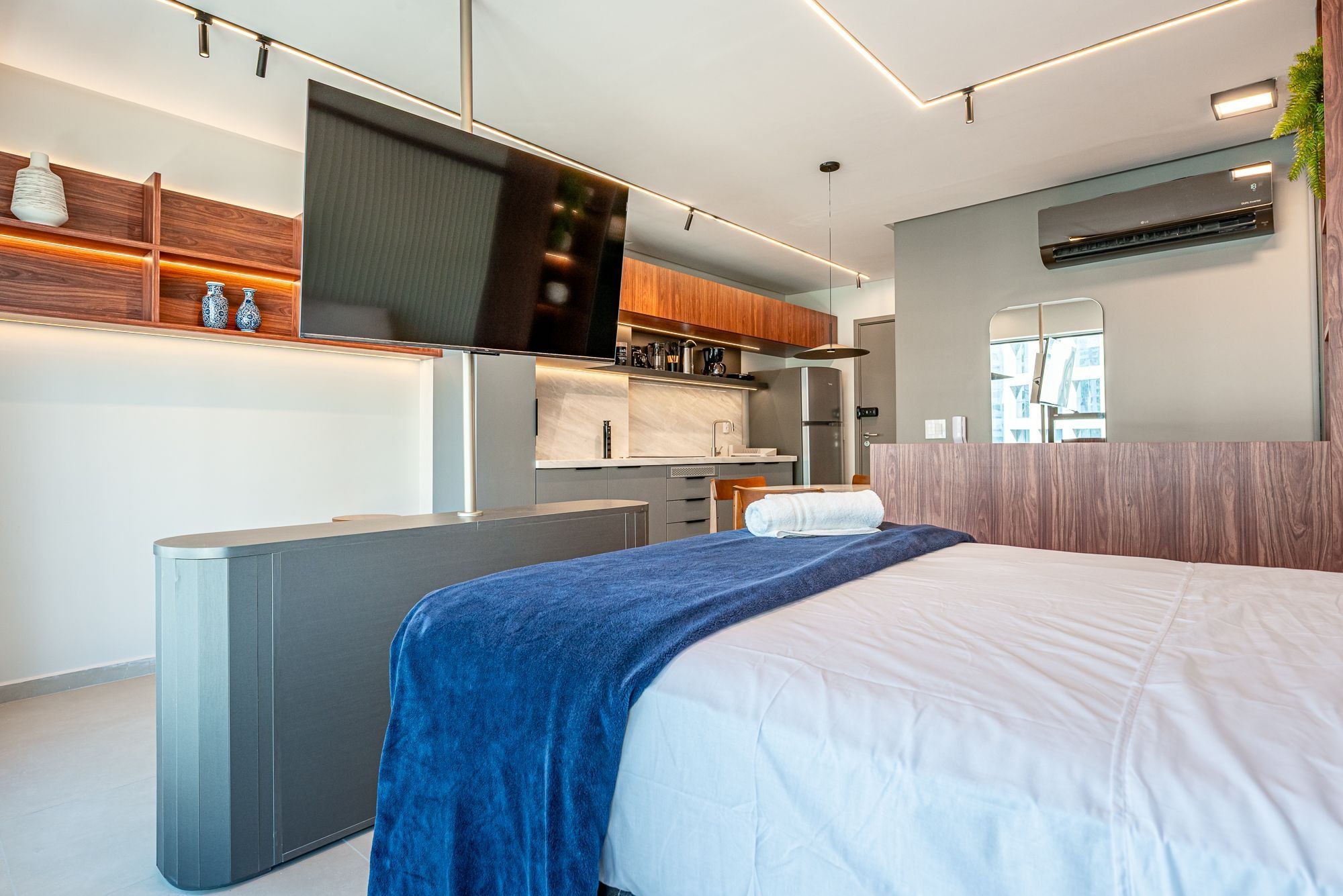 A modern hotel room with a bed, a desk, a kitchenette, and a large window.