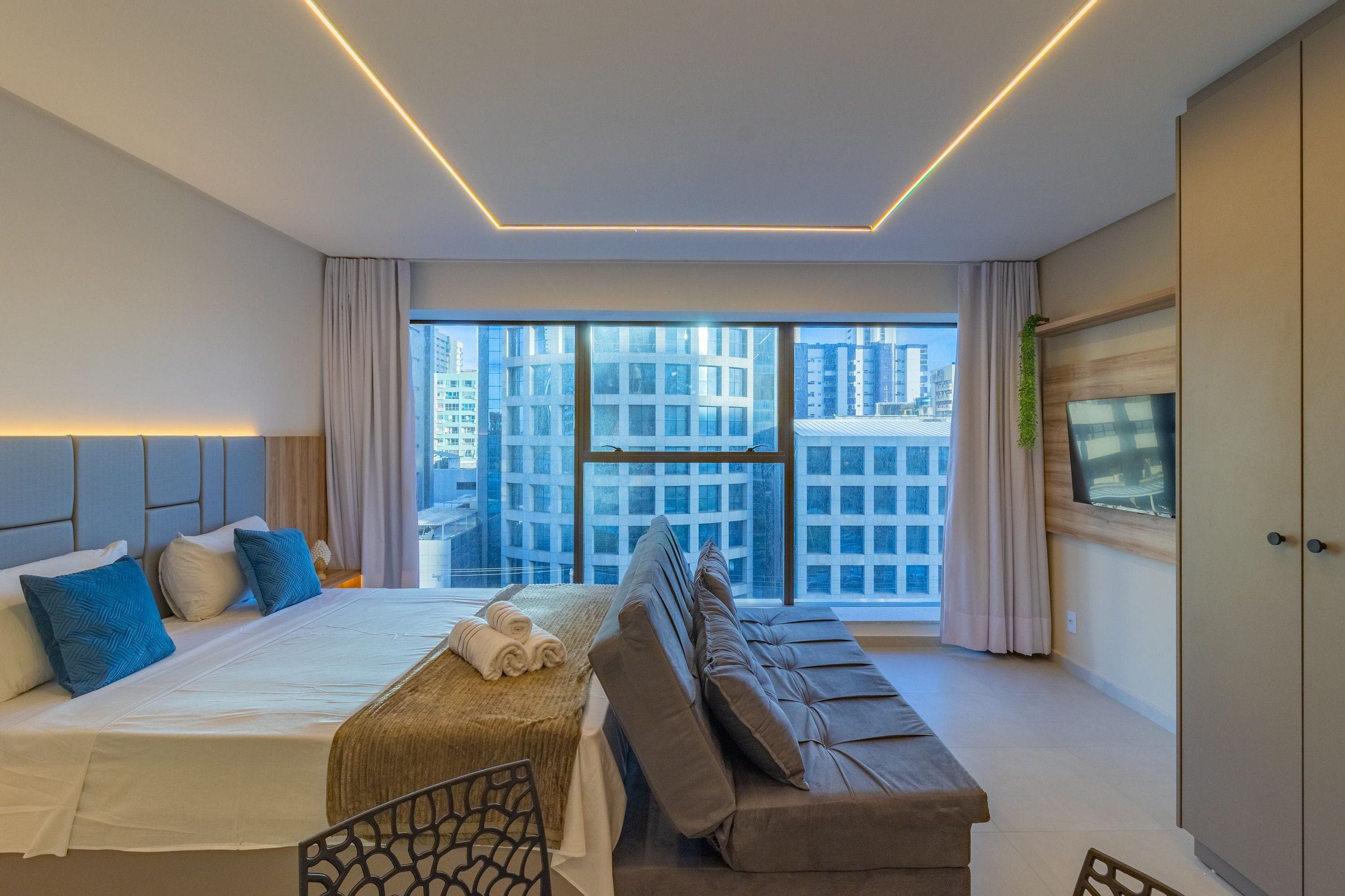 Quarto de hotel moderno com uma cama grande, um sofá e uma TV. O quarto possui janelas amplas com vista para a cidade.