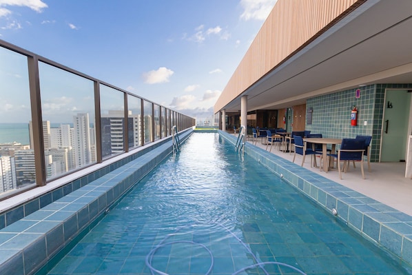 Une piscine à débordement située sur le toit, offrant une vue sur la ville, un espace détente avec des tables et des chaises, et un sol carrelé.