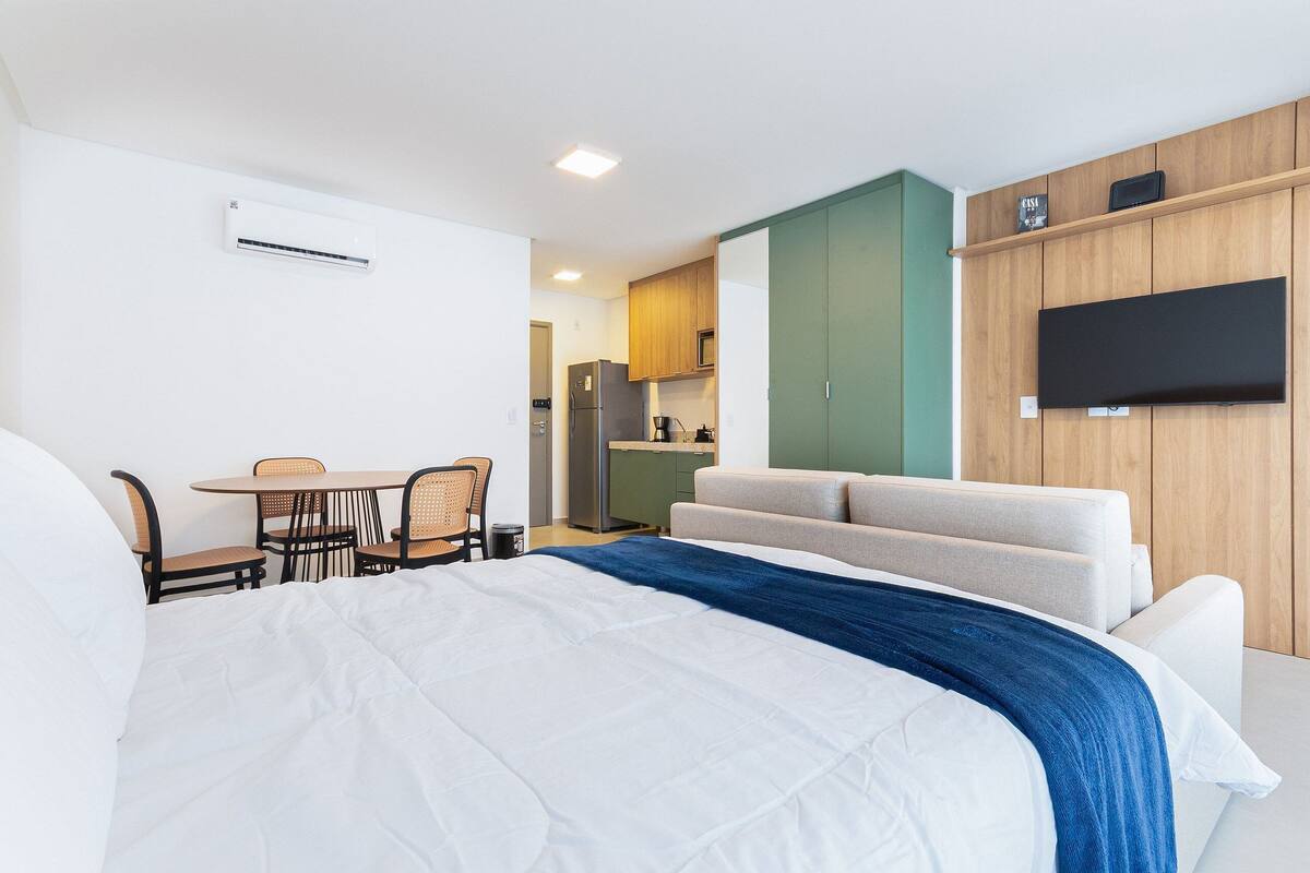 A hotel room with a large bed, a sofa, a dining table with chairs, a kitchen area, and a flat-screen TV mounted on the wall.