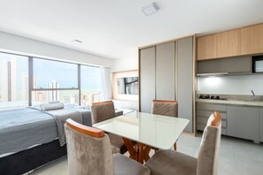 Een moderne hotelkamer met een bed, zithoek, eettafel, keuken en grote ramen met uitzicht op de stad.