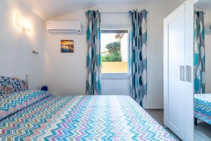 Un dormitori amb un llit, una finestra amb cortines, un aire condicionat fixat a la paret i una fotografia petita.