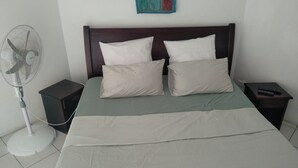 Un letto con testiera in legno, due cuscini bianchi, una coperta verde, una ventola portatile e un comodino con un telecomando.