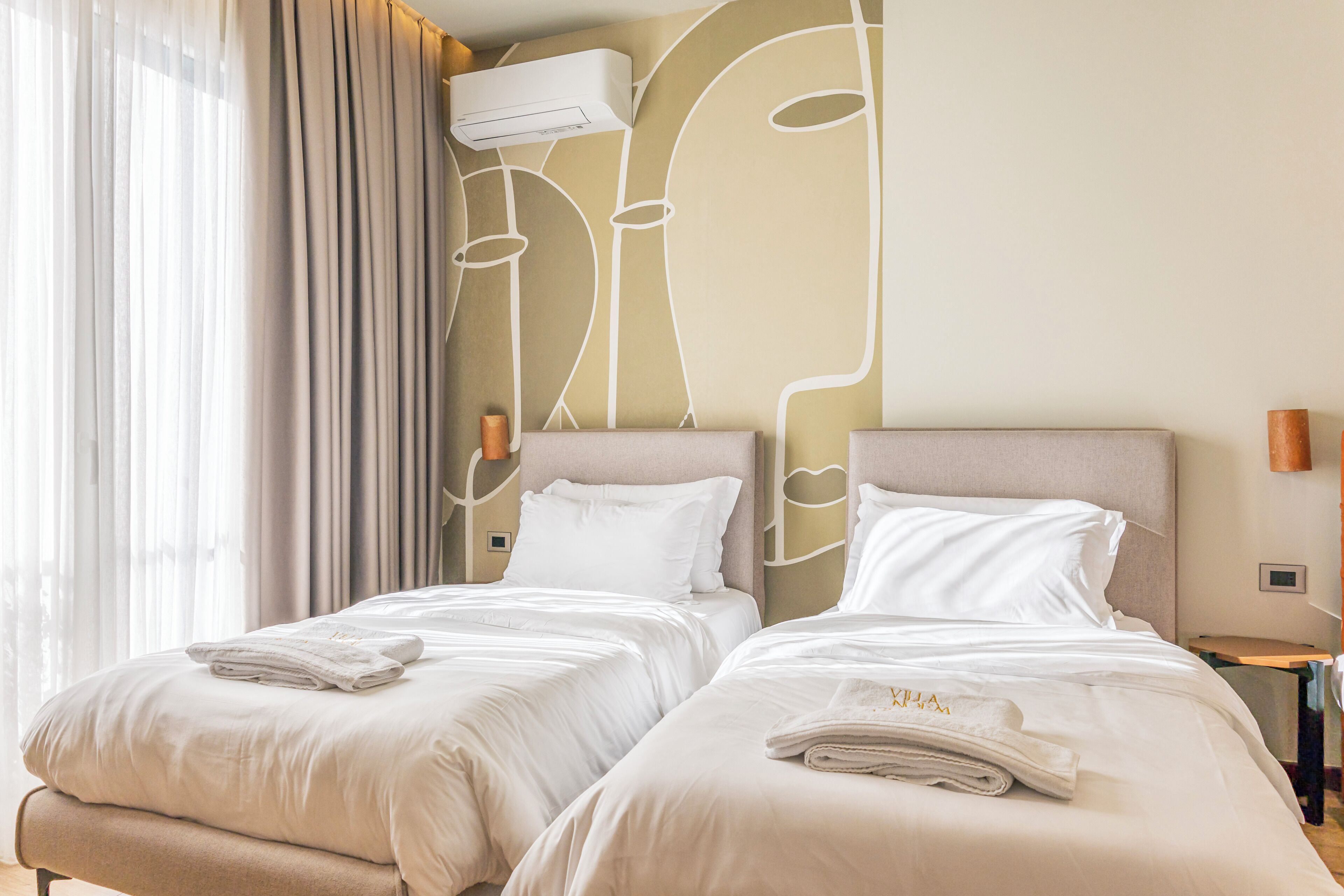 Ein Hotelzimmer mit zwei Betten, einem großen Fenster mit Vorhängen, einer Klimaanlage und einer Wand mit abstraktem Design.
