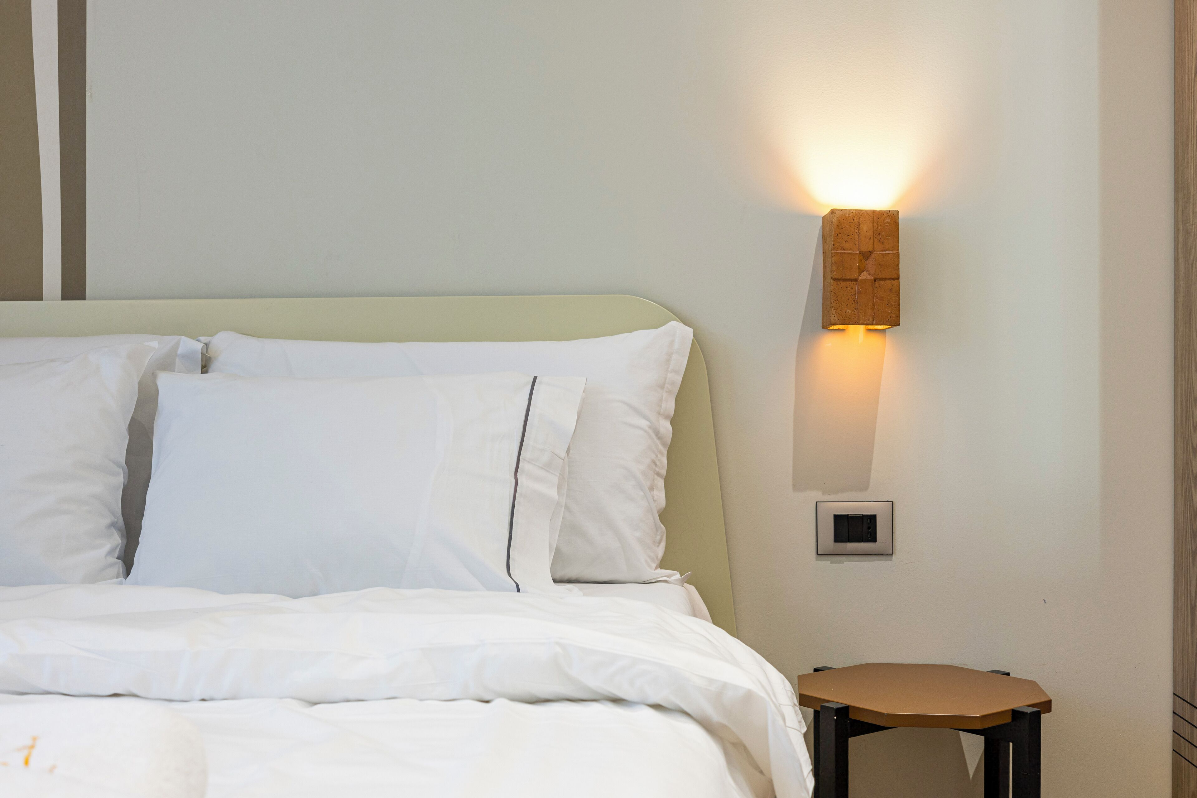 Una cama bien hecha con sábanas blancas, un taburete de madera y una lámpara fijada a la pared.