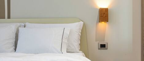 Una cama bien hecha con sábanas blancas, un taburete de madera y una lámpara fijada a la pared.