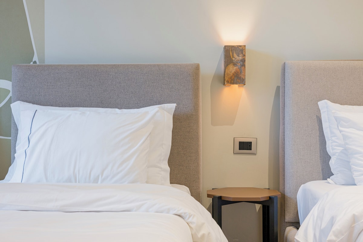 Habitación de hotel con dos camas, cada una con ropa blanca y cabeceros en tonos neutros. Hay una mesita de noche pequeña con una lámpara y un aplique de pared.