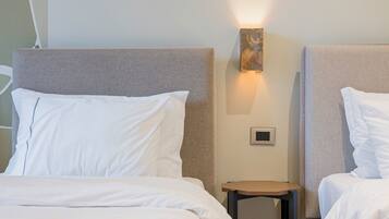 Une chambre d’hôtel avec deux lits, chacun doté de draps blancs et d’une tête de lit aux couleurs neutres. On y trouve une petite table de chevet avec une lampe, ainsi qu’un luminaire fixé au mur.