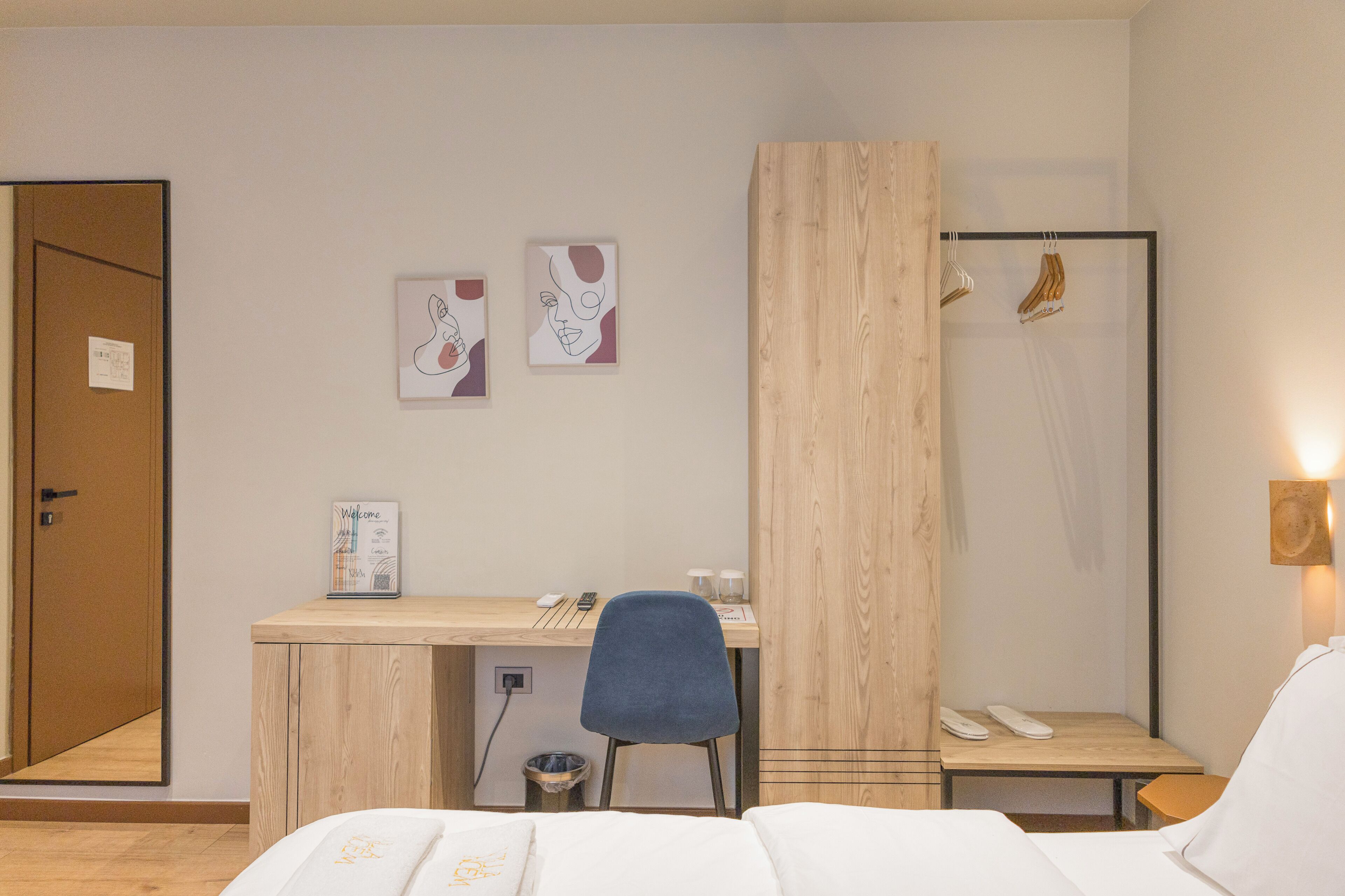 Hotelkamer met een houten bureau, een stoel, een bed met witte beddengoed, een kleine prullenbak en een houten garderobe.