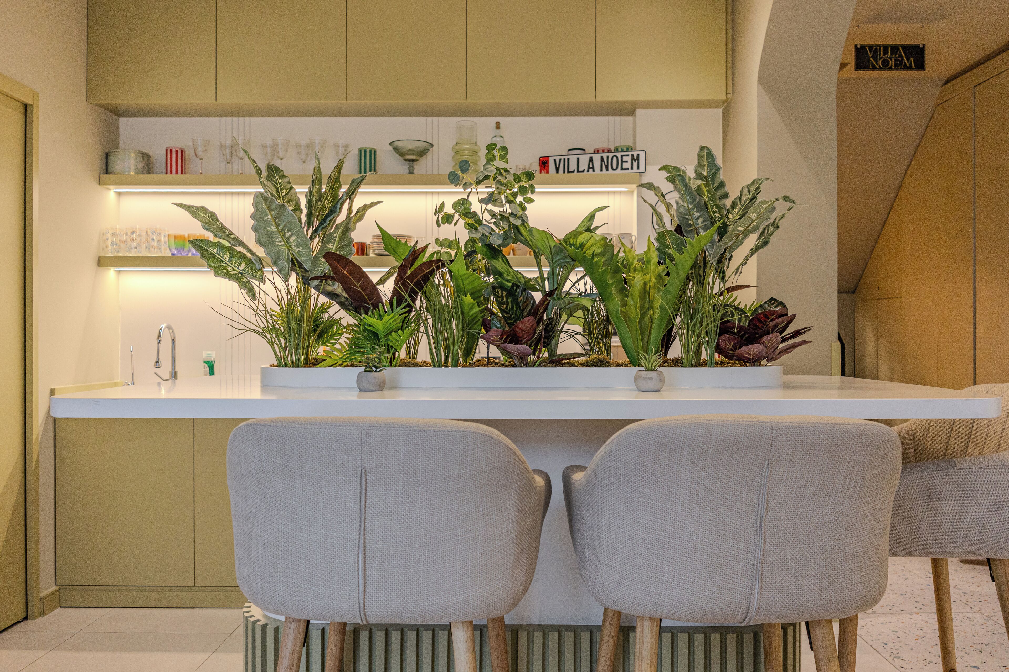 Cozinha moderna com bancada branca, duas cadeiras cinzas e diversos vasos com plantas.