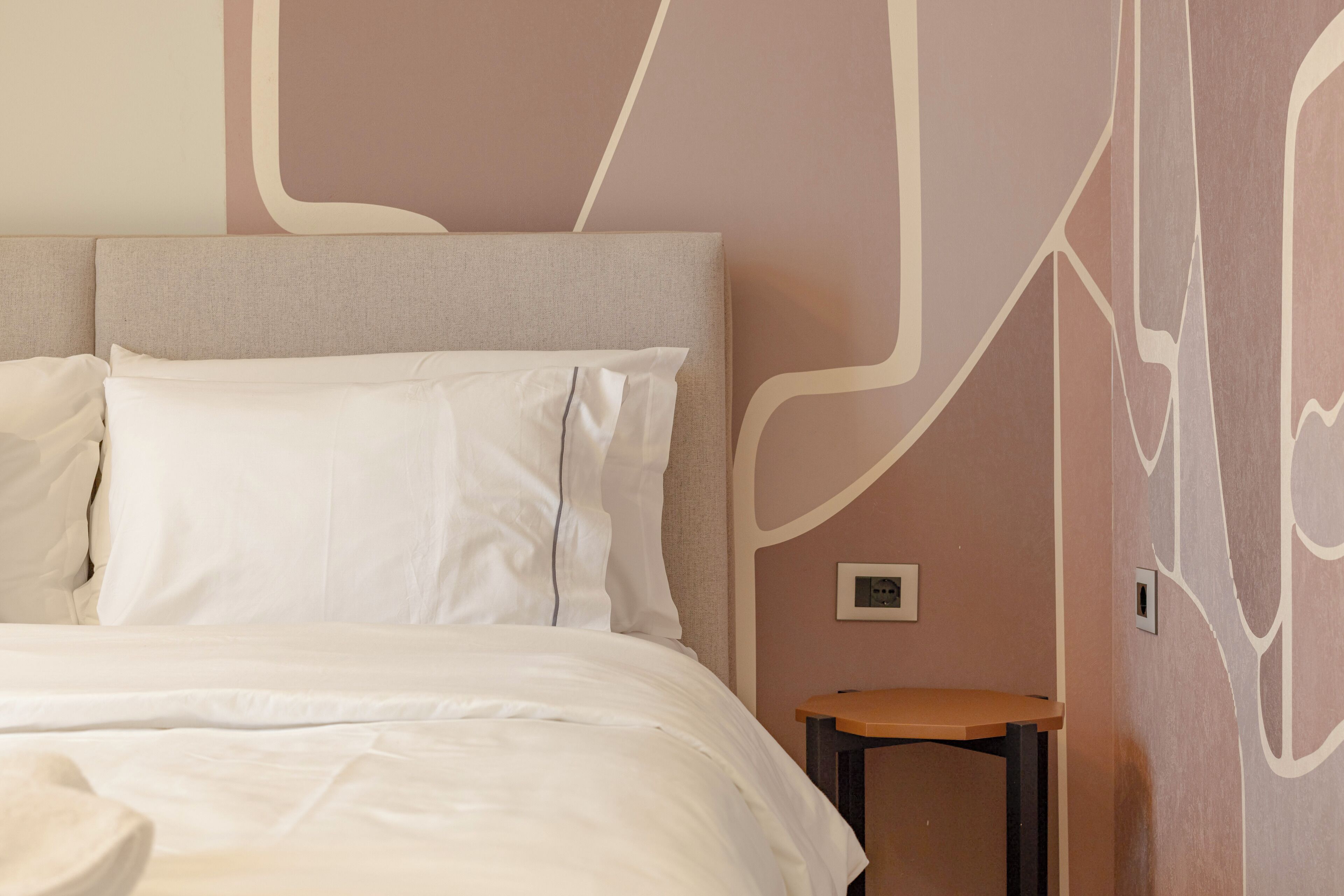 Una cama bien hecha con sábanas blancas en una habitación de diseño moderno, incluyendo un cabecero y una pared con motivos abstractos.