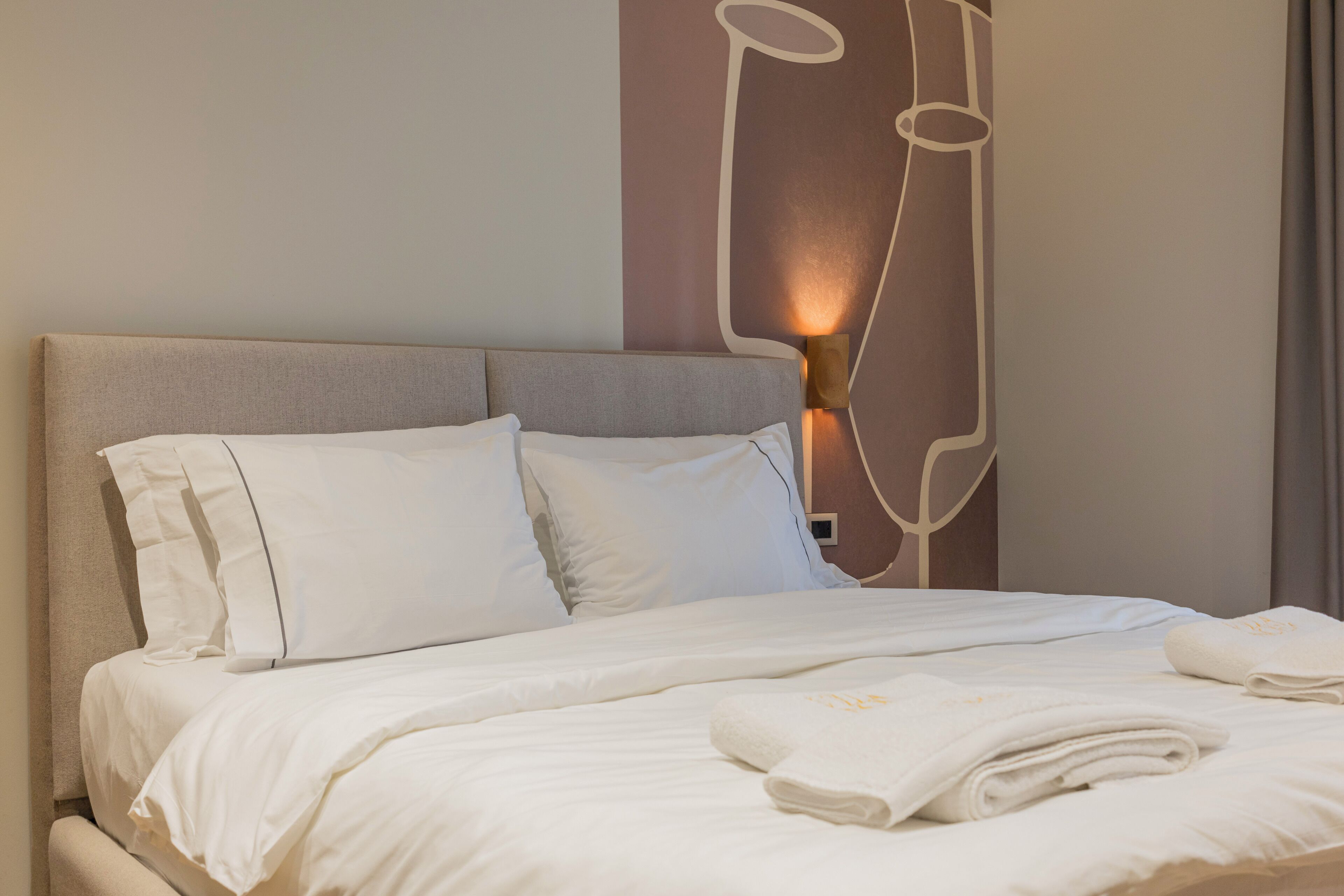 Un lit bien fait, avec du linge de lit blanc et des oreillers, un luminaire fixé au mur et une fenêtre avec des rideaux.