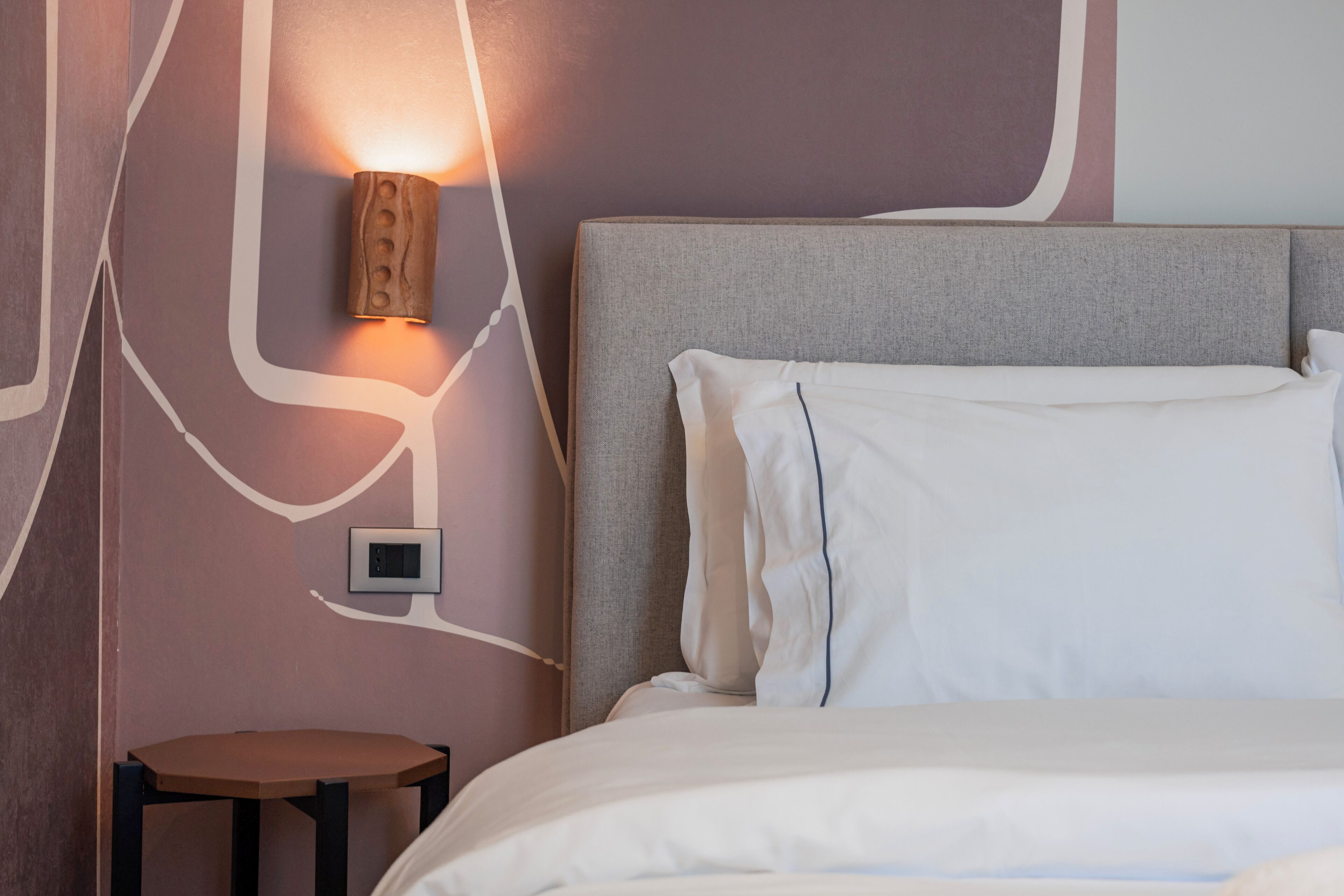 A modern hotel room with a grey headboard, white bedding, a small bedside table, and a wall-mounted light fixture.