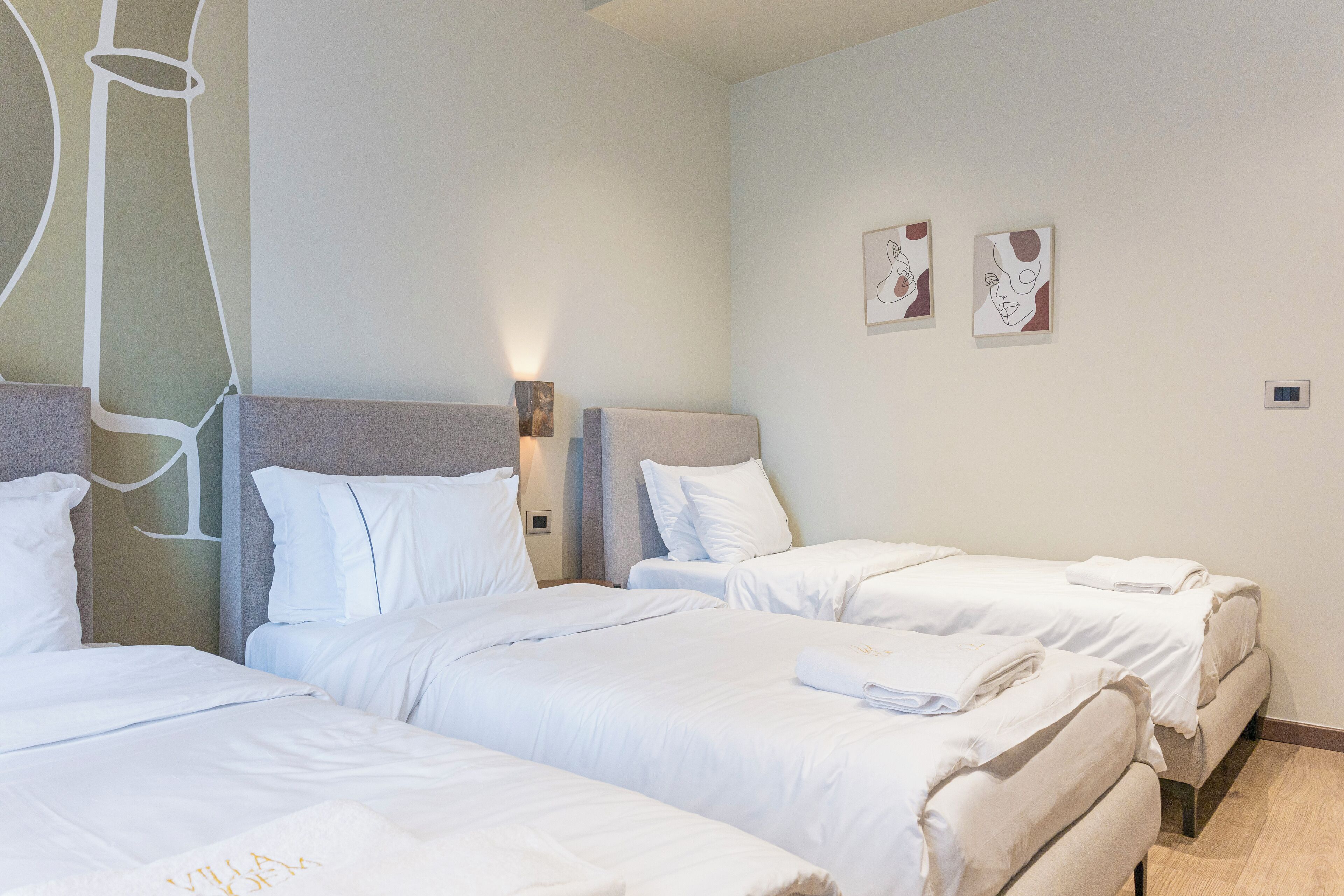A hotel room with three beds, each with white bedding and pillows. There are two wall-mounted artworks and a small bedside table with a lamp.