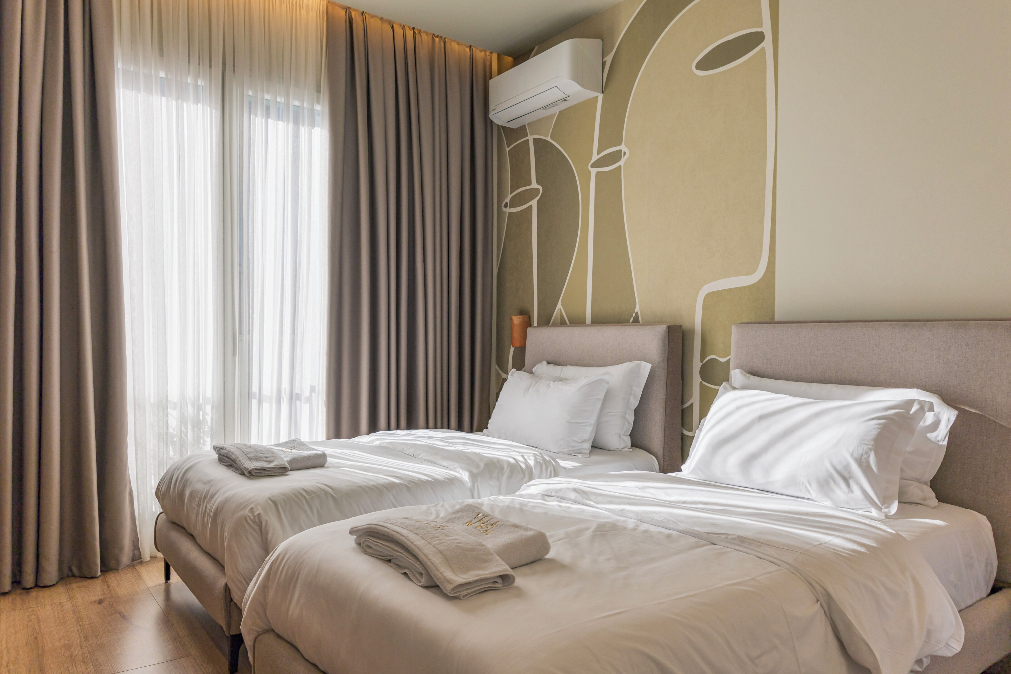 A hotel room with two beds, a large window with curtains, and an air conditioning unit on the wall.