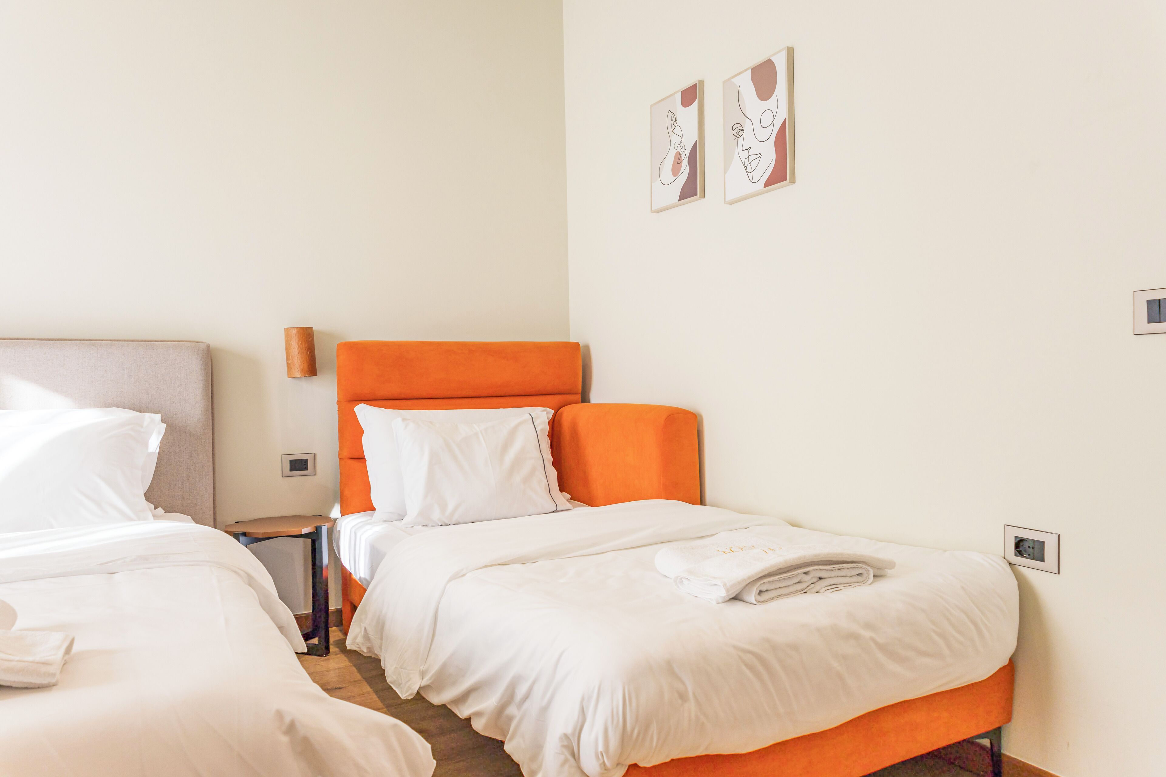 A hotel room with two beds, one with an orange headboard, white bedding, and a neatly folded towel on the bed. There are two wall-mounted light switches and two framed artworks on the wall.